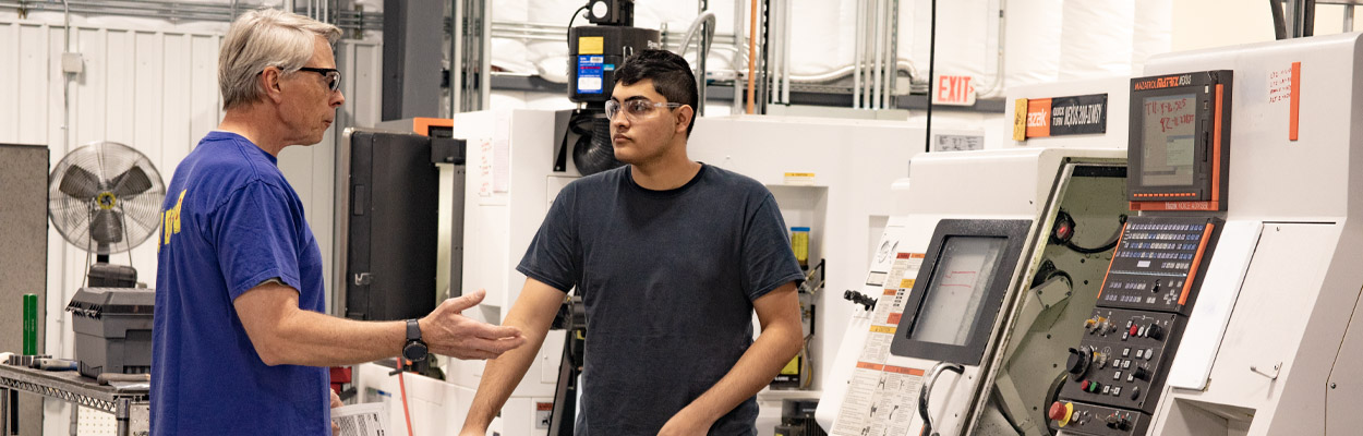 An older machinist instructs a younger machinist in using a CNC machine.